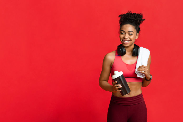 Happy african fitness girl with towel on her shoulder and wireless headset holding bottle with protein, looking at copy space